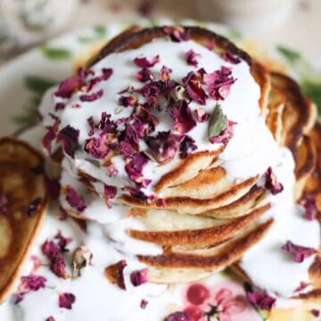 A stack of pancakes topped with white icing and sprinkled with dried rose petals is presented on a floral-patterned plate. The background is slightly blurred, showcasing a soft, cozy atmosphere.