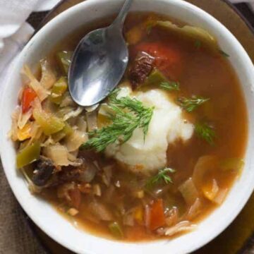 A bowl of vegetable soup with a dollop of sour cream is garnished with dill. A spoon rests in the bowl, ready to raise a spoonful of goodness, and a white cloth with red stripes is beside it on a brown surface. The soup contains visible chunks of vegetables such as carrots and celery—this is one of the good soups.