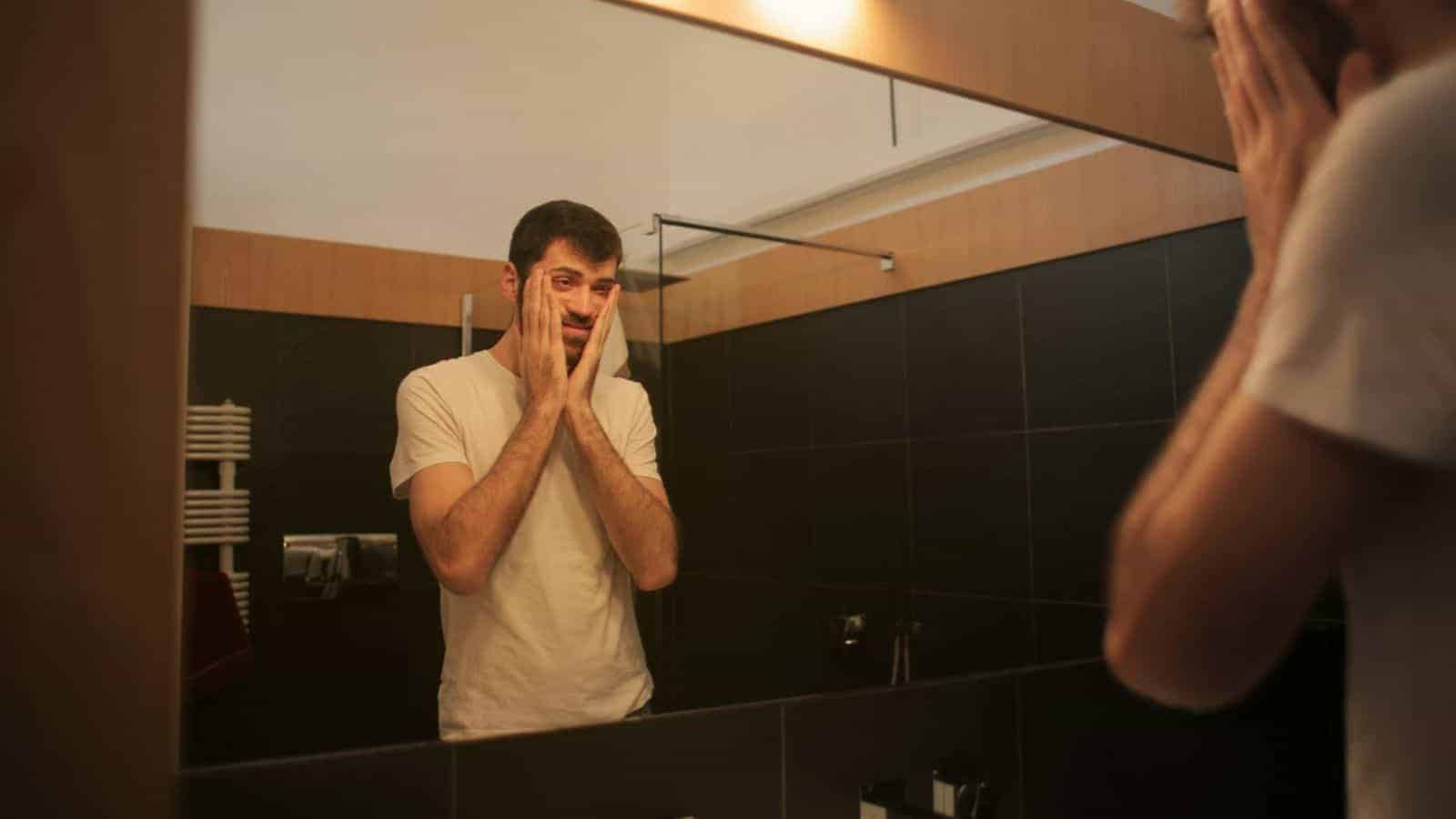 A man in a white t-shirt is standing in front of a bathroom mirror with a concerned expression, holding his face with both hands. The bathroom has dark tiles and over-sink lighting.