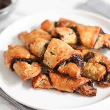 A white plate holds several pieces of golden-brown baked rugelach with a visible filling of dark chocolate or fruit spread. A bowl with a similar dark substance is faintly visible in the background. The plate rests on a light gray fabric.