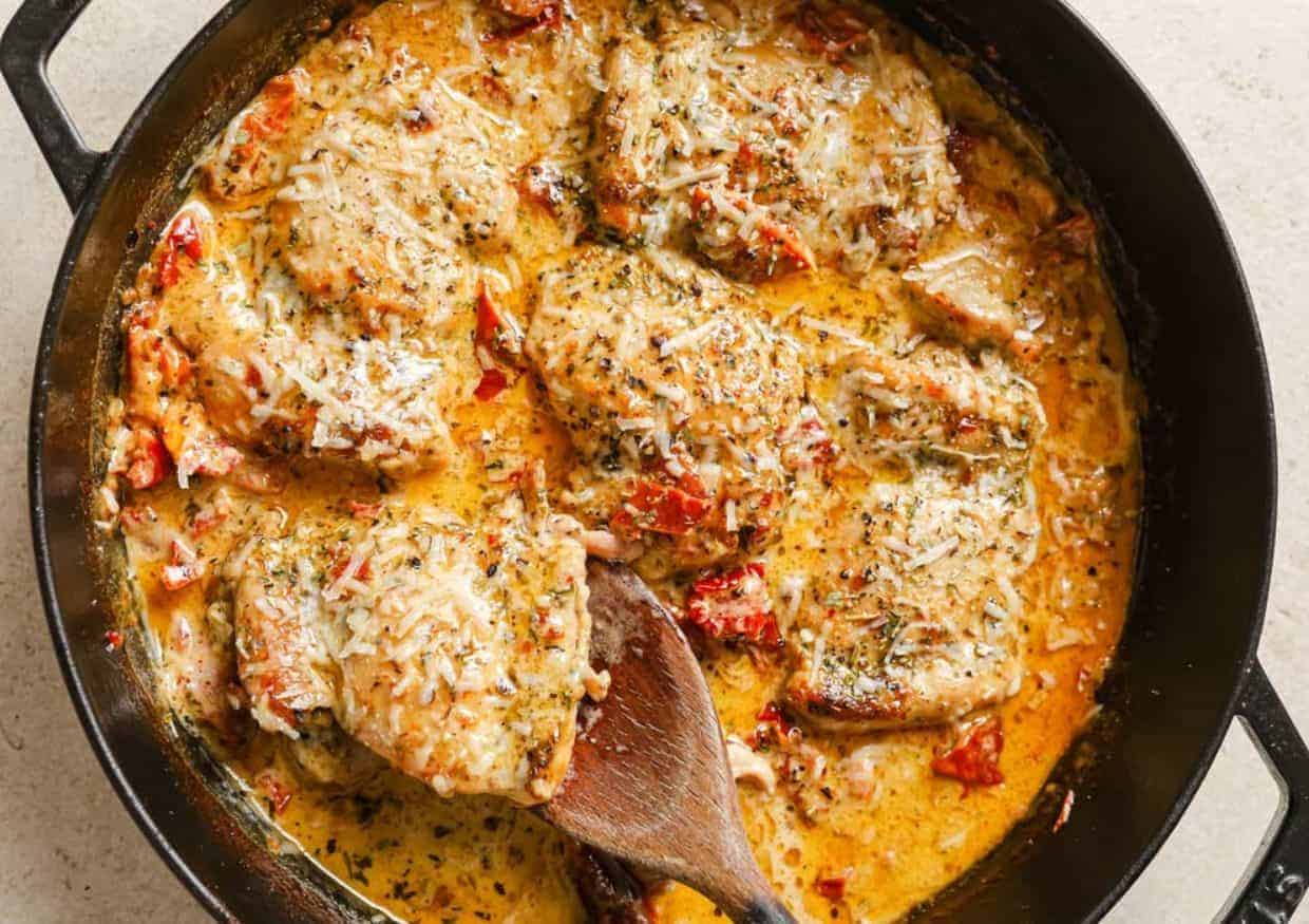 A skillet filled with cooked chicken breasts in a creamy tomato sauce, topped with shredded cheese and herbs—a perfect addition to your weeknight dinner recipes. A wooden spoon rests in the sauce, and pieces of sun-dried tomatoes are visible throughout the dish.