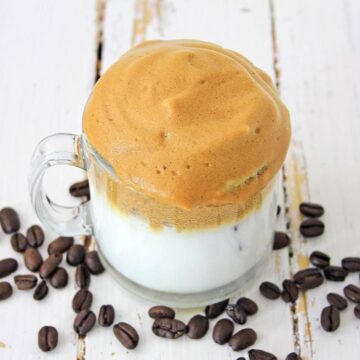 A glass mug filled with a white milk base topped with a thick layer of frothy, whipped coffee sits on a rustic white wooden surface. Coffee beans are scattered around the mug.