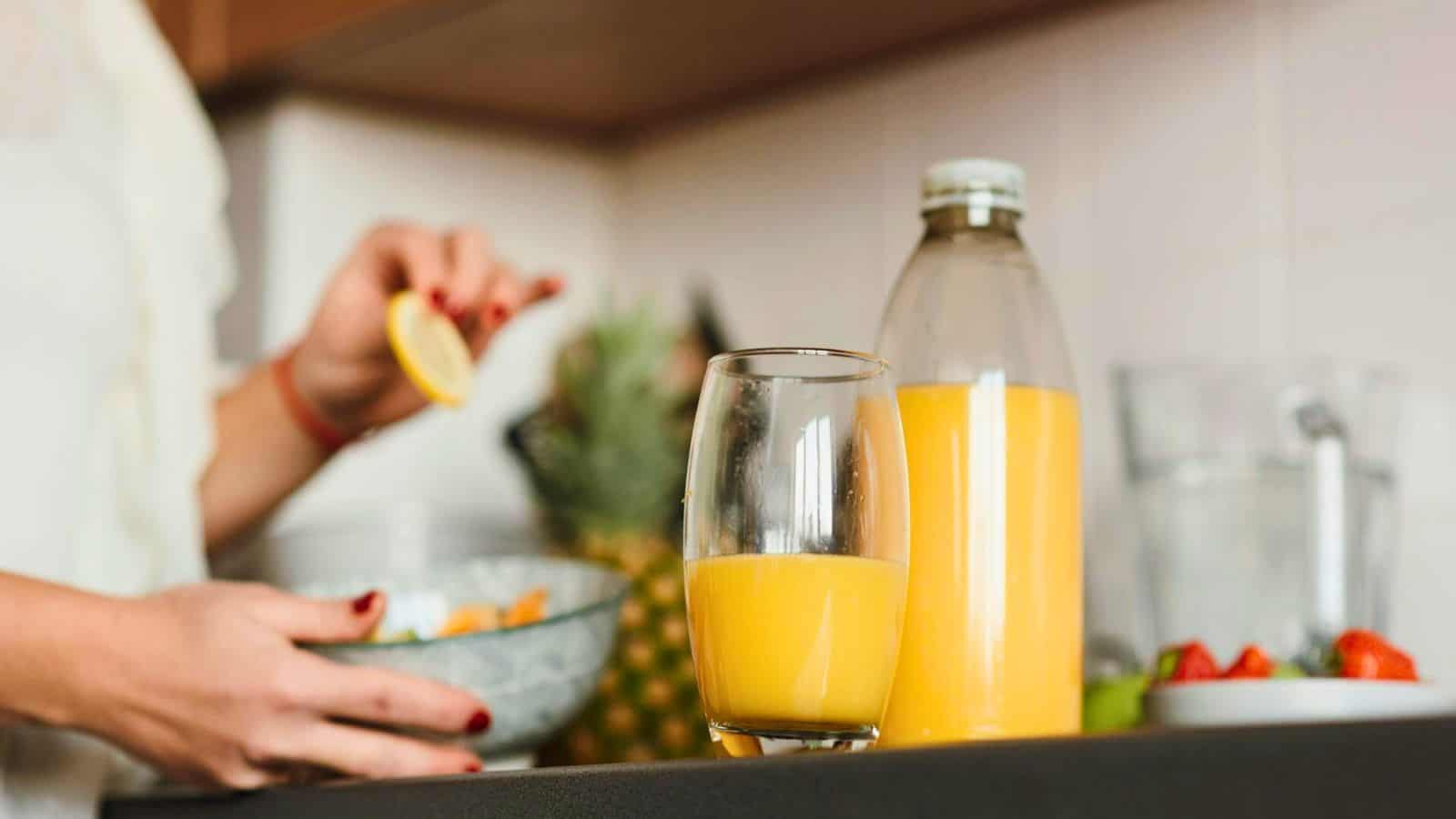 A person is preparing food in a kitchen, holding a lemon slice over a bowl of salad. In the foreground, there's a glass and a bottle of orange juice on the counter. Various fruits, including strawberries and a pineapple, are visible in the background. The scene busts food myths about healthy eating being bland.