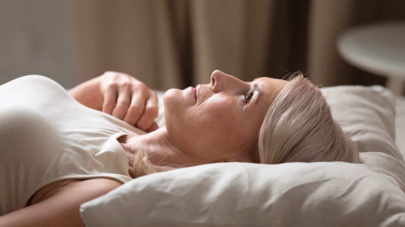 A woman with blonde hair is lying on a pillow, gazing upward. She is wearing a white sleeveless top. The background is out of focus, consisting of beige curtains and a round table. The setting appears to be a calm, indoor environment.