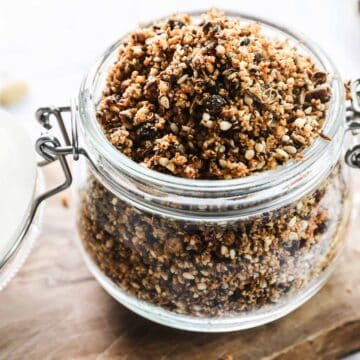 A glass jar filled with granola sits on a wooden cutting board. The jar is open, with the lid resting beside it. The granola, reminiscent of a dukkah recipe, is mixed with various seeds, nuts, and dried fruit. A wooden spoon is partially visible next to the jar.