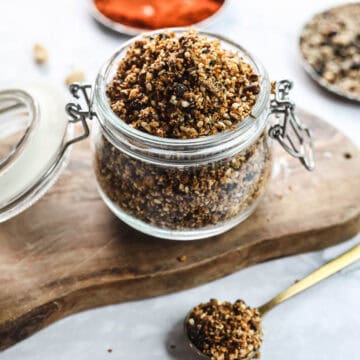 A glass jar filled with granola is placed on a wooden surface. The jar has a metal clasp, and its lid is open. A spoonful of granola lies beside the jar. In the background, scattered nuts and a bowl containing Egyptian-style dukkah add an exotic touch to this authentic recipe setup.