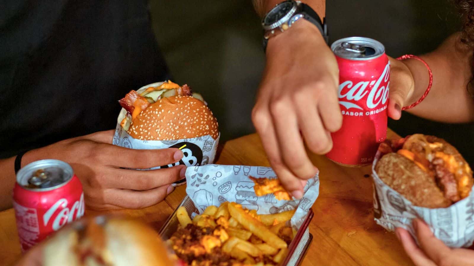 Two people are sitting at a table eating fast food. One person holds a cheeseburger, while the other reaches for crinkle-cut fries. Both have open cans of Coca-Cola and one has a sandwich. The wooden table is covered with food wrappers, defying food myths about healthier options.