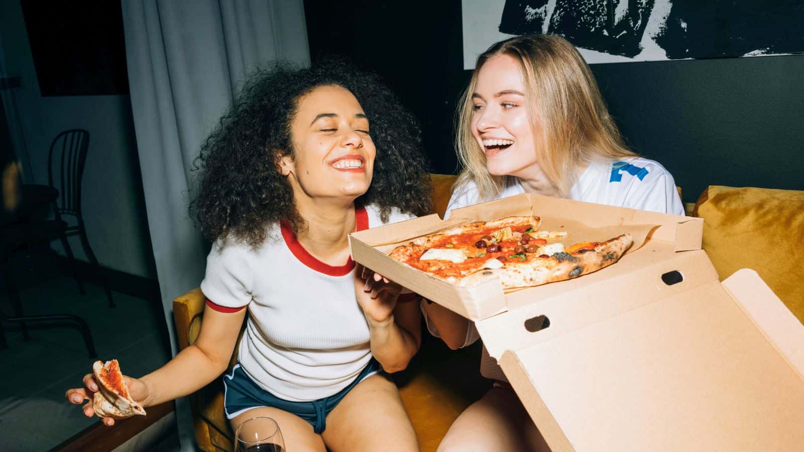 Two women are sitting on a yellow couch. One woman is holding an open pizza box with a pizza inside, and the other woman is holding a slice of pizza. Both are smiling and appear to be enjoying their time while debunking food myths. There is a chair and a curtain in the background.