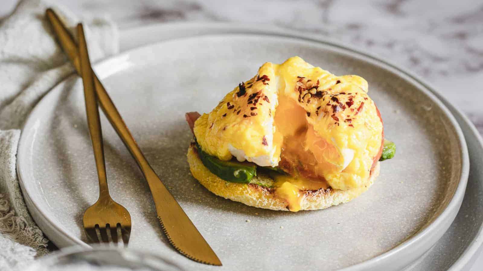 A plated dish features an English muffin topped with avocado, bacon, and a poached egg covered in hollandaise sauce. The yolk is partially split, showing the running yolk, debunking common food myths about eggs. A gold fork and knife are set next to the plate. The background includes a textured white cloth.