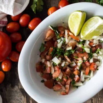 A white bowl filled with chopped tomatoes, onions, cilantro, and drizzled with lime juice is placed on a wooden table, reminiscent of iconic Colombian dishes found in Bogotá. Two lime wedges sit beside the bowl. Cherry tomatoes and cilantro are scattered next to a white cloth with red stripes.