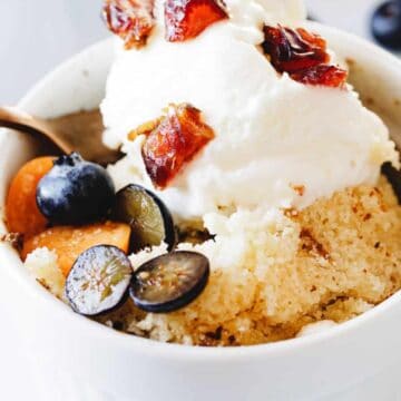 A white bowl contains a serving of couscous topped with whipped cream, chopped dates, blueberries, and slices of another fruit, possibly figs or plums. A spoon is partially visible in the bowl, with a few blueberries scattered around, creating an alluring tableau of dessert classics.