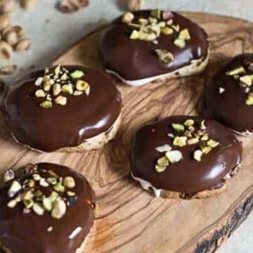 This image shows a wooden tray with five chocolate-coated pastries topped with crushed nuts, possibly pistachios. The tray, seemingly placed as though by auto draft, is on a light-colored surface, and scattered nuts are visible around the tray.