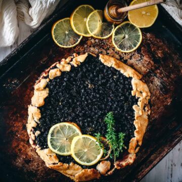 A rustic berry galette topped with slices of lemon and a sprig of thyme is displayed on a weathered baking sheet. Several lemon slices and a honey dipper with honey are placed at the top-right corner of the sheet. A white cloth is partially visible on the left.