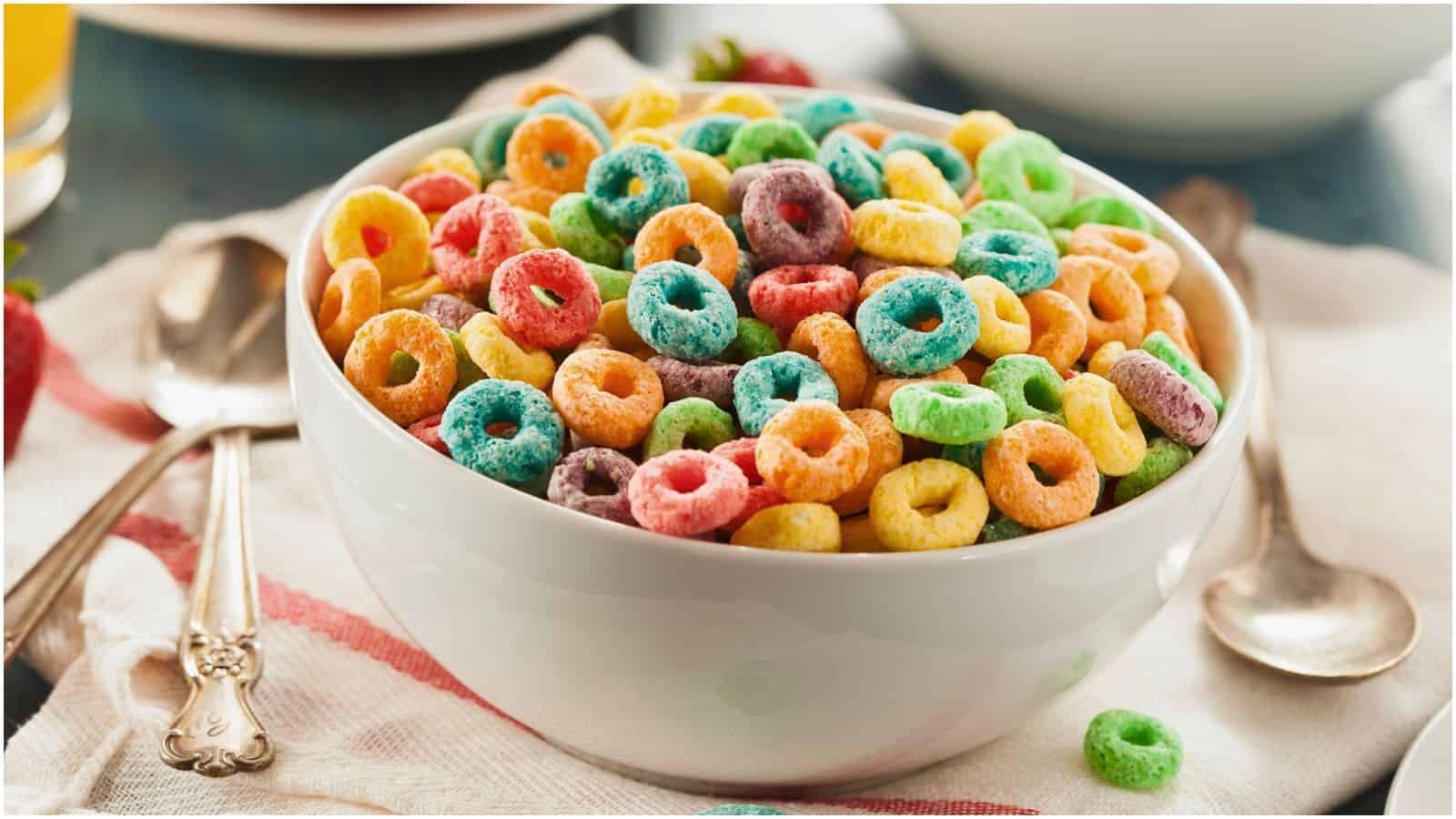 A white bowl filled with multi-colored, ring-shaped cereal sits on a white cloth with a spoon nearby. The cereal includes colors such as red, orange, yellow, green, blue, and purple. A second spoon is also visible, along with a plate and a glass of orange juice.