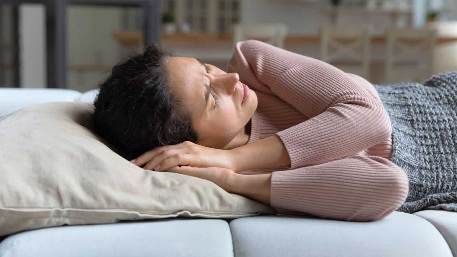 A woman lies on her side on a couch, resting her head on a pillow. She is wearing a pink sweater and appears to be looking off into the distance with a contemplative expression. In the background is a kitchen area with chairs around a table.