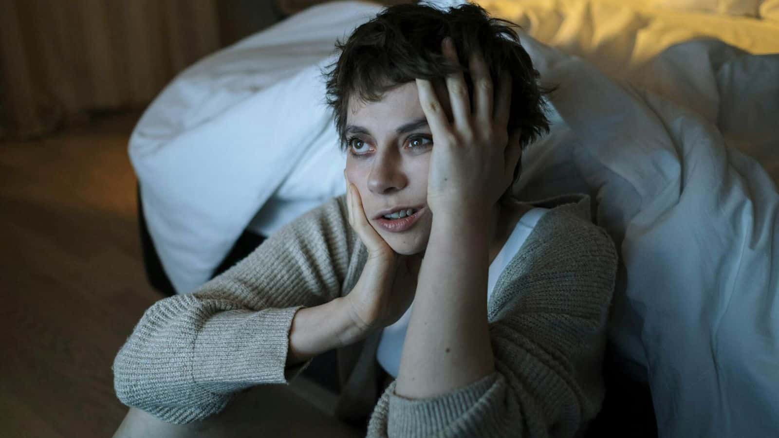 A person with short hair is sitting on the floor beside a bed, holding their head with both hands and looking up with a thoughtful expression. The room appears dimly lit, with disheveled bedding in the background.