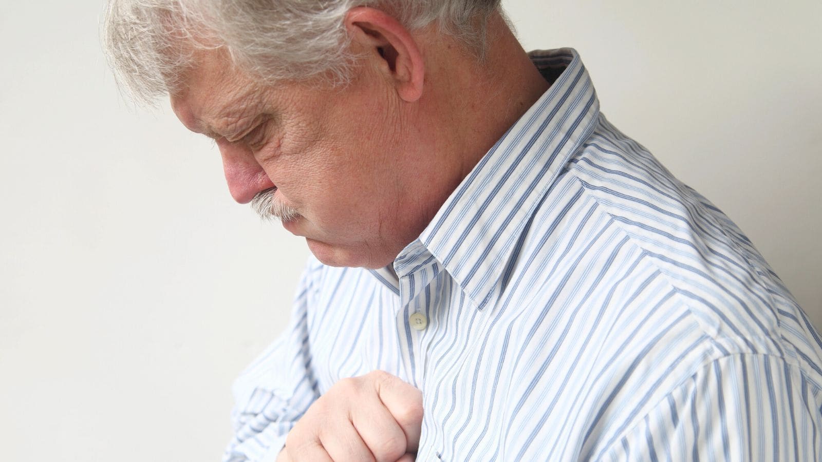 An elderly man with grey hair and a mustache is looking down with his hand placed on his chest. He is wearing a blue and white striped shirt. The background is a plain off-white wall.