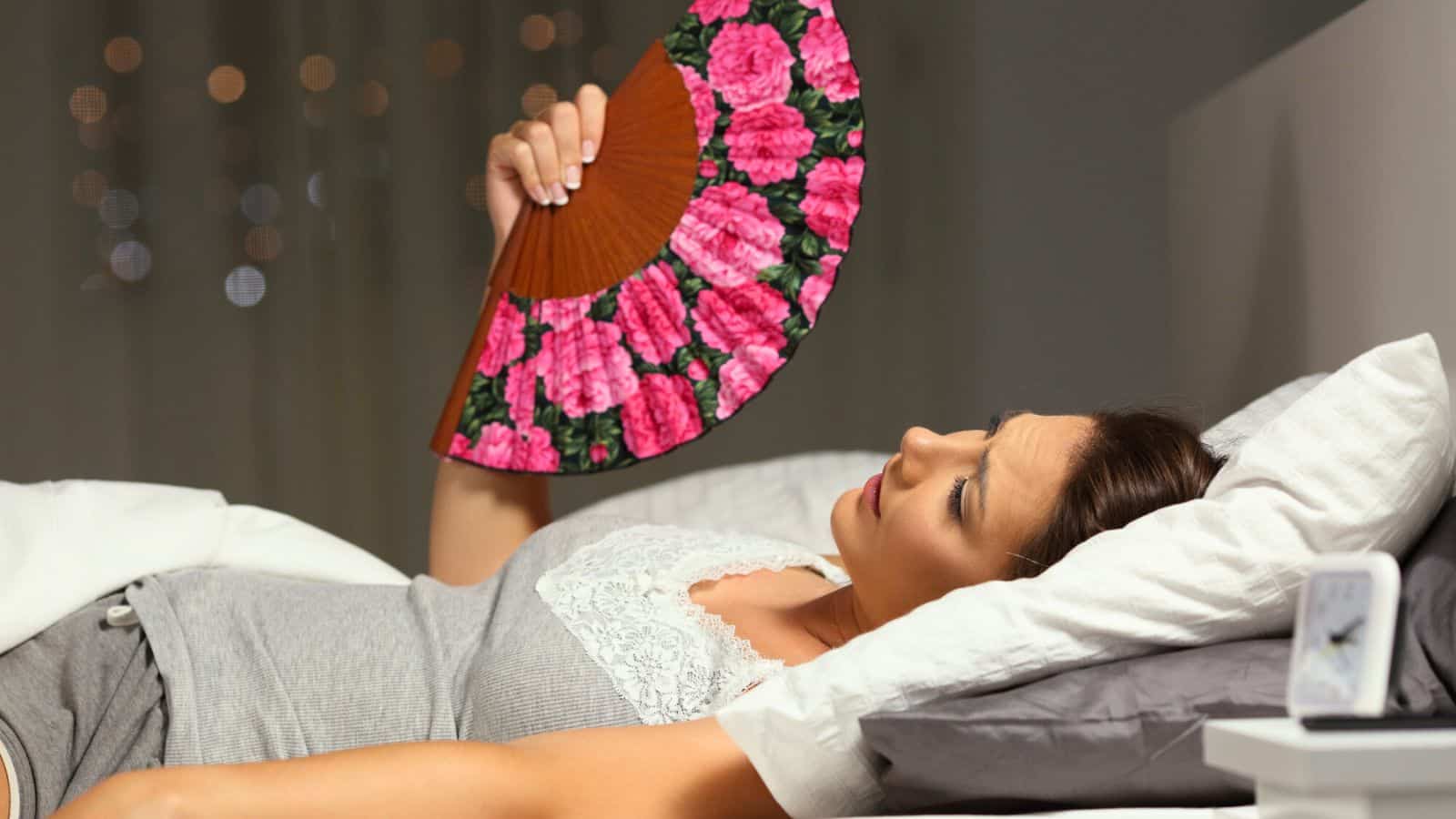 A woman lies on a bed covered with a white blanket, holding a floral hand fan above her head. She wears a grey tank top and grey shorts. The background features blurred lights and the corner of a bedside table with a white digital clock.