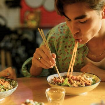 A person wearing a floral shirt is seated at a wooden table, using chopsticks to eat noodles from a bowl. The individual is leaning over the bowl, slurping the noodles. Another bowl of food and a hand holding chopsticks are visible in the foreground.