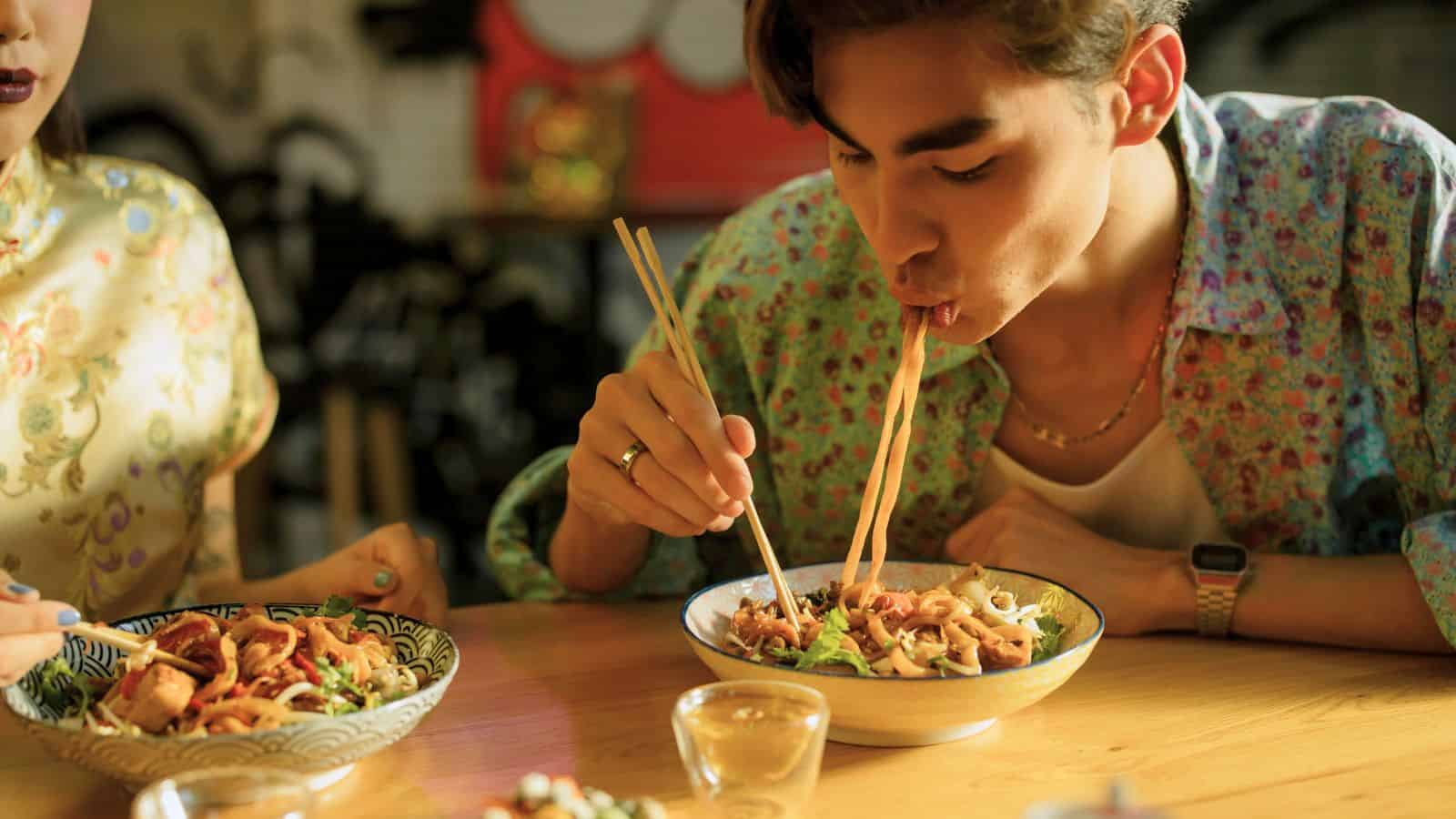 A person wearing a floral shirt is seated at a wooden table, using chopsticks to eat noodles from a bowl. The individual is leaning over the bowl, slurping the noodles. Another bowl of food and a hand holding chopsticks are visible in the foreground.