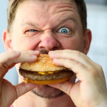 A man is holding a hamburger with both hands close to his mouth. With a playful expression, one eye squinting and the other wide open, he seems ready to debunk food myths. Dressed in a pink shirt, the background behind him is blurred.