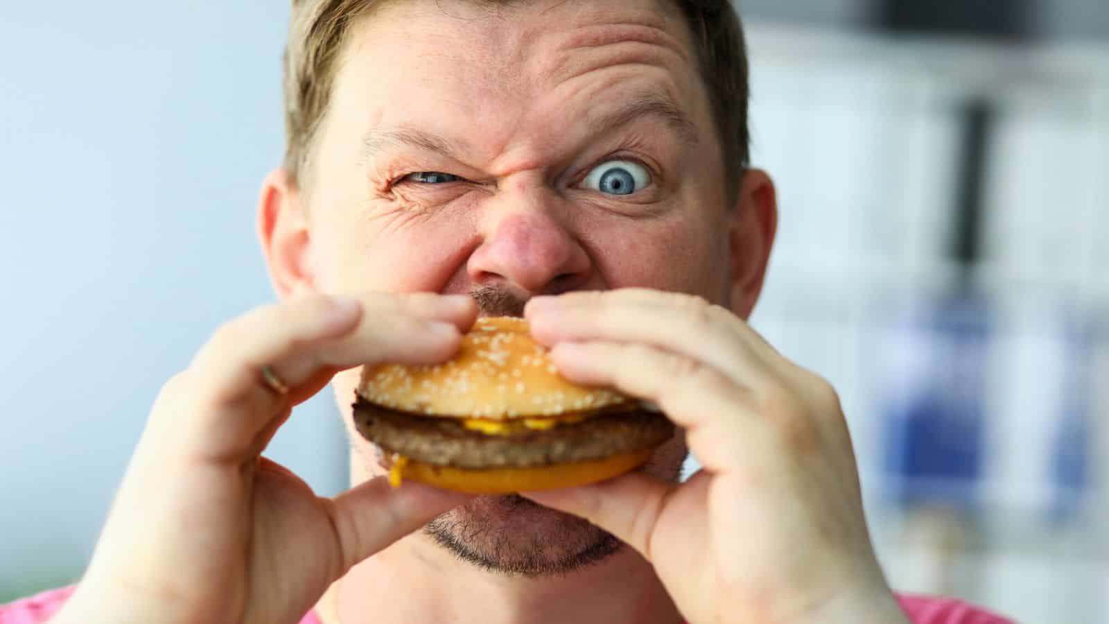 A man is holding a hamburger with both hands close to his mouth. With a playful expression, one eye squinting and the other wide open, he seems ready to debunk food myths. Dressed in a pink shirt, the background behind him is blurred.