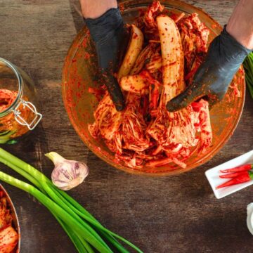 Person wearing black gloves is mixing kimchi in a large bowl. Ingredients including jars of kimchi, green onions, garlic, chili peppers, and spices are on a wooden table. A small bowl of red pepper flakes and a dish of coarse salt are also present, perfect for anyone fermenting for beginners.