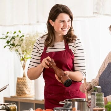 Two women in a kitchen, smiling as they cook together and share old school kitchen tips. The younger woman, wearing a red apron, is using a pepper grinder, while the older woman, in a light blue apron, stirs a pot. The counters are adorned with various ingredients and utensils.