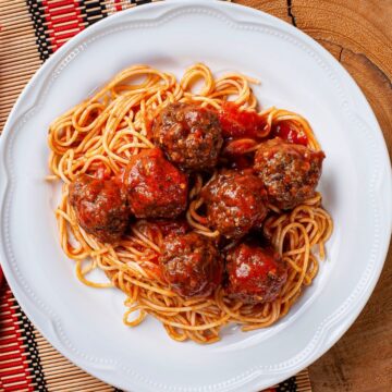 A white plate filled with spaghetti and meatballs in tomato sauce is placed on a wooden surface with a woven placemat. A fork, knife, and red napkin are positioned to the left side of the plate, embodying perfect cooking pasta tips.