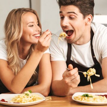A woman and a man, both wearing white shirts and black aprons, are sitting at a kitchen counter eating different types of noodles from plates. The woman is feeding the man a forkful of spaghetti while both are smiling. There are salt and pepper shakers on the counter.