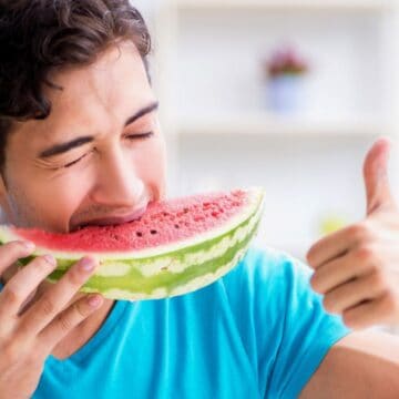 A man in a blue shirt is enjoying a large slice of hydrating watermelon, his eyes closed and smiling as he takes a bite. His right hand gives a thumbs-up gesture. The background, blurred with light-colored shelves, emphasizes his delight in one of nature’s most refreshing fruits and veggies.