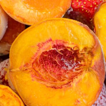 Close-up of halved peaches, showcasing the bright orange and yellow flesh with the pits removed. The peaches have a slightly frosty appearance, suggesting they are frozen. A few strawberries are also visible in the background.