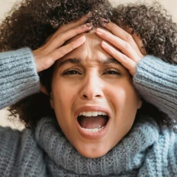 A person with curly hair is wearing a light blue sweater and holding their head with both hands. They have a distressed expression with their mouth open, as if they've just heard one of those things people who don't drink alcohol are tired of hearing. Light beige background.