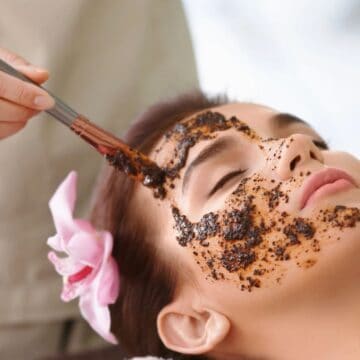 A person is lying down with eyes closed while another applies a face mask using a brush. The mask, dark brown and granular, seems to incorporate coffee in your beauty routine. The individual receiving the treatment has a pink flower in their hair, enhancing the serene atmosphere.
