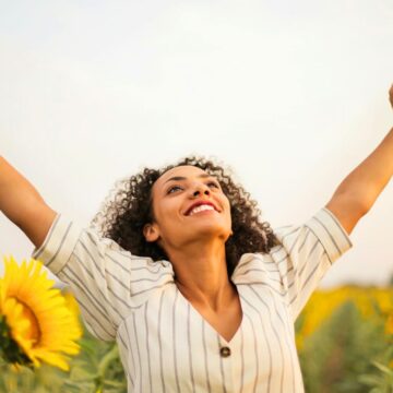 A woman stands in a sunflower field with her arms raised and a joyful expression on her face, embracing the essence of self-care tips. She is wearing a striped blouse and has curly hair. The sky is clear, and sunflowers are visible in the background.
