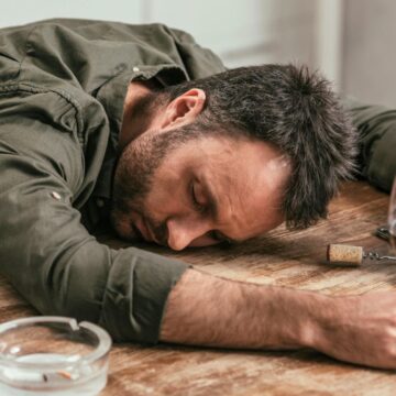 A man with dark hair and a beard is asleep on a wooden table, his head resting on his arm. He holds a wine glass with red wine in one hand and a wine bottle in the other, showing early signs of alcoholism. An ashtray, a cork, and a corkscrew are also on the table.