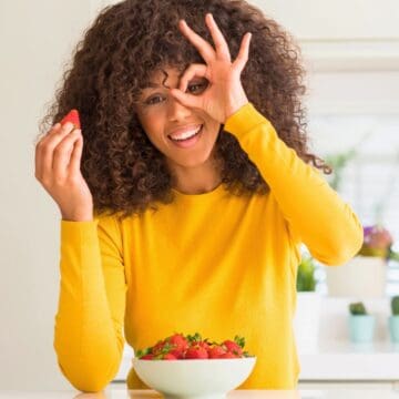 A person with curly hair, wearing a yellow shirt, is holding a strawberry in one hand and making an "OK" gesture with the other hand over their eye. There is a bowl of strawberries on the table in front of them. The background includes some potted plants.