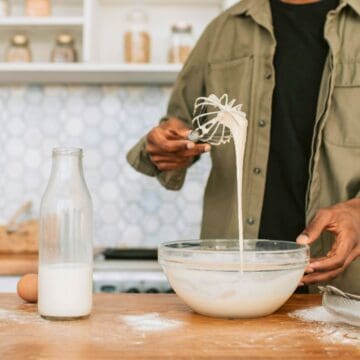 A person stands in a kitchen, whisking batter in a bowl. Nearby on the counter are a jar of milk, a glass bottle of milk, an egg, a whisk, and a few blueberries. The background shows a backsplash with a geometric pattern and kitchen shelves with jars.
