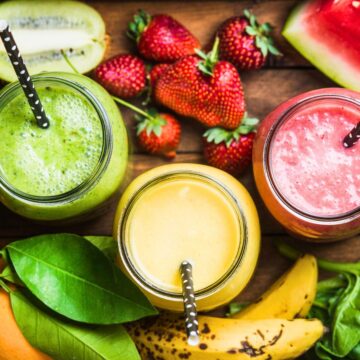 Three glasses of healthy jamba juice drinks with black and white polka dot straws are placed on a wooden table surrounded by fruits and leafy greens. From left to right, the smoothies are green, yellow, and pink. Fruits include kiwi, strawberries, banana, and watermelon.