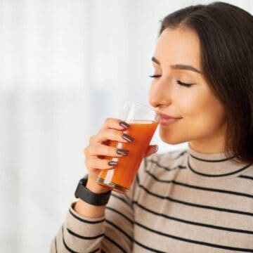 A woman with long dark hair is holding a glass of orange-colored juice close to her lips, appearing to enjoy the drink. She is wearing a long-sleeved, beige and black striped shirt, and a smartwatch. The background is softly blurred with white curtains, evoking an image of someone savoring drinks for high blood pressure.