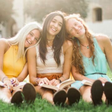 Three young women sit on grass in a park, leaning against each other and smiling. They are holding open books, suggesting they are reading or studying together. Trees and a building are visible in the background on a sunny day.