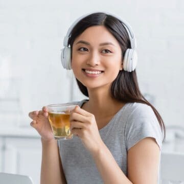 A woman with long hair wearing white headphones and a grey t-shirt is holding a glass cup with tea. She is smiling and sitting at a table in a bright kitchen with white brick walls and shelves in the background.