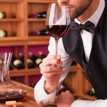 A person in formal attire, with a bow tie and vest, smells a glass of red wine in a wine cellar. Bottles of wine are visible on wooden racks in the background, and a decanter with red wine sits on a wooden table nearby along with a couple of corks.