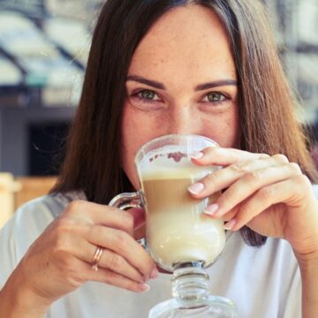 A woman with long brown hair is sitting indoors, holding a clear glass mug and drinking a latte. She is looking directly at the camera with a neutral expression. She is wearing a white shirt, and the background is blurry with indoor lighting.