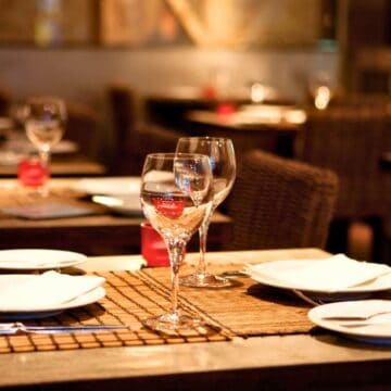 A restaurant dining area with several wooden tables and wicker chairs. Each table is set with white plates, wine glasses, cutlery, and napkins on brown woven placemats. The room is softly lit, creating a warm and inviting atmosphere.