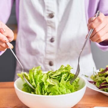 A person in a white apron is tossing a green salad with two forks in a white bowl on a wooden tabletop. Another plate with salad is visible beside the bowl. The background includes a kitchen counter and cabinets.