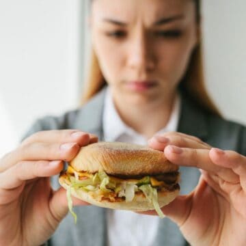 A person in a gray blazer is holding a sandwich with both hands and looking at it with a displeased expression. The sandwich contains lettuce and other visible ingredients. The background is plain white.