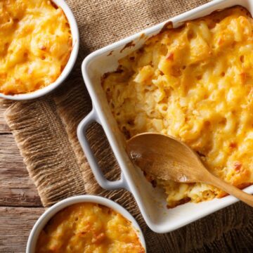 A rectangular white baking dish filled with baked macaroni and cheese is placed on a piece of burlap fabric on a wooden table. Two smaller round dishes also containing macaroni and cheese are beside the main dish. A wooden spoon rests in the main dish.