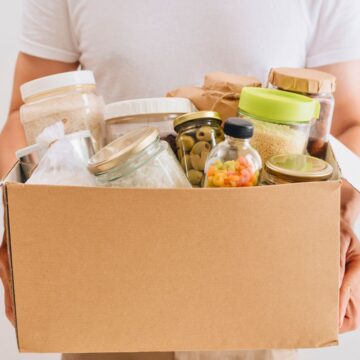 A person is holding a cardboard box filled with various jars and containers, showcasing some of the best foods to stockpile. These containers appear to hold different types of food items, such as pasta, grains, and olives. The person is wearing a white t-shirt, and the background is plain white.