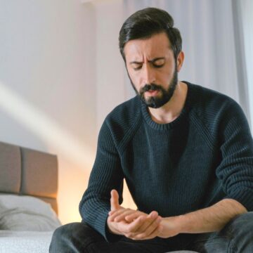 A bearded man with short dark hair and wearing a dark sweater is sitting on a bed in a brightly lit room. He is looking down at his hands with a concerned expression. In the background, there are pillows and a headboard. The room has white curtains.